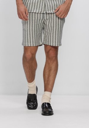 Man draagt zwarte leren loafers, crèmekleurige sokken en geruite grijs-witte knielange shorts tegen een effen achtergrond.