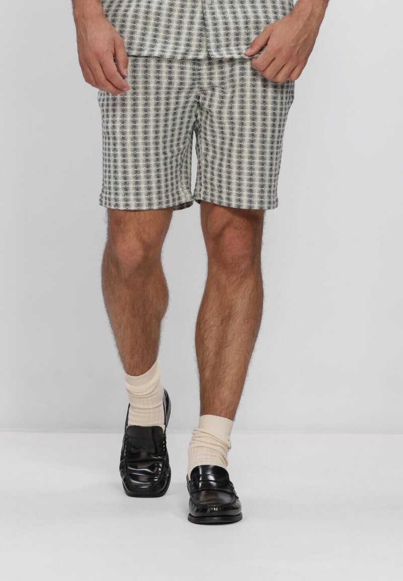 Homme portant des mocassins en cuir noirs, des chaussettes crème et un short à carreaux gris et blanc jusqu'aux genoux sur un fond uni.