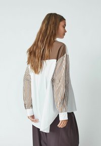 Maglietta a maniche lunghe con un design a blocchi di colore marrone e bianco, maniche a righe e orlo curvo. Il tessuto sembra liscio e leggero.