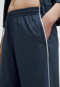 Tamno plave atletske hlače od glatkog materijala. Imaju elastični pojas s vezicom i bijele bočne pruge. Uključuju bočni džep.