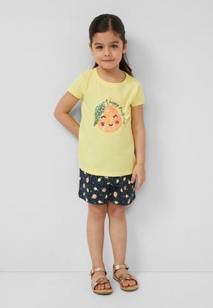 Meisje met donker haar dat een geel T-shirt met een glimlachende oranje afbeelding en marineblauwe korte broek met een fruitpatroon draagt, staand tegen een effen achtergrond.