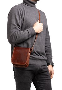 Borsa a tracolla in pelle marrone con forma quadrata, cuciture chiare a contrasto e tracolla regolabile, indossata a spalla.