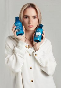 Två blå schampoflaskor märkta "Redken Extreme" hålls i en persons händer, som bär en vit skjorta med knappar och ett pärlhalsband.