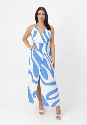 Vrouw staat met hand op heup en draagt een blauwe en witte halterjurk met abstracte print en witte open-toe hakken.