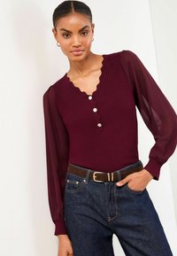 Burgunderfarbener gerippter Body mit wellenförmigem Ausschnitt und Knopfdetails, kombiniert mit dunkelblauen Denim-Jeans und einem braunen Gürtel, der durchsichtige Puffärmel aufweist.
