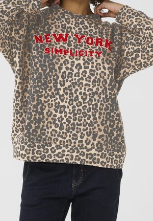 Sweat à imprimé léopard avec texte rouge "NEW YORK SIMPLICITY" porté par une personne ajustant le col, associé à un jean bleu foncé.