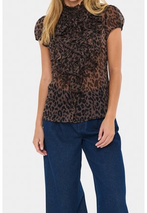 Vrouw draagt een bruine blouse met luipaardprint met ruches aan de voorkant en korte pofmouwen, gecombineerd met donkerblauwe jeans met wijde pijpen.