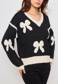Pull en maille noir avec des accents crème et des motifs de nœud. Design en V avec un ourlet et des poignets côtelés. Texture douce avec une coupe confortable.