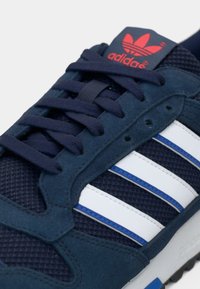 Tenisky z modrého semiše a síťoviny s bílými a modrými pruhy, kulatými tkaničkami a červeným logem na jazyku. Mají texturovaný svršek.