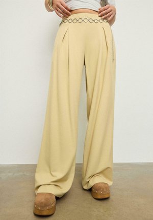 Béžové kalhoty se širokými nohavicemi a předními záhyby, nošené se stříbrným řetízkovým páskem a hnědými dřeváky na hladkém podkladu.