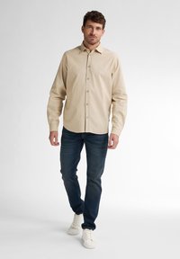Beige knopenlijnshirt van lichtgewicht stof, met lange mouwen en een gladde textuur. Gecombineerd met donkerblauwe jeans en witte sneakers.