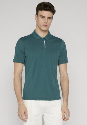 Ung mann iført en turkis kortermet polotrøye med glidelås og hvite shorts, stående mot en ensfarget lys bakgrunn.