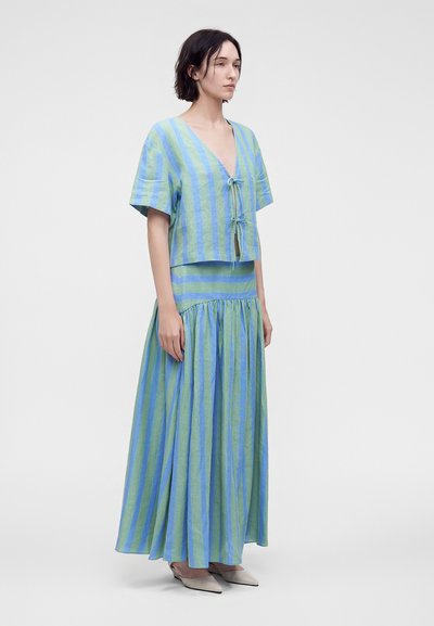 Femme portant un haut ample rayé bleu et vert à col en V avec liens et une longue jupe fluide assortie, sur un fond clair uni.