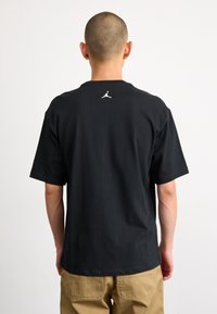 Černé bavlněné tričko s krátkými rukávy a kulatým výstřihem. Na zadní straně se nachází bílé logo Jordan. Hladká textura, volný střih.