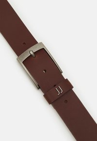 Brązowy skórzany pasek z srebrnym metalowym klamrą, o eleganckim designie, z widocznymi otworami do regulacji i tłoczonym detalem w pobliżu klamry.