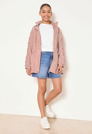 Young person smiling, wearing a light pink jacket, white shirt, denim shorts, and white sneakers, standing against a plain white background.