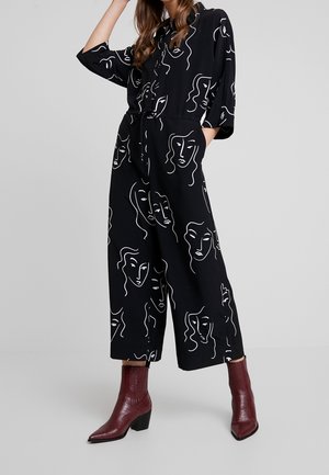 Femme portant une combinaison noire à jambes larges avec un motif abstrait de visage en lignes blanches et des bottines à talons bordeaux, debout avec une main dans la poche.