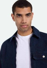 Tengerészkék farmerdzseki cipzárral és mellzsebbel. Belső, fehér póló látható. Puha tapintású és strukturált gallér.