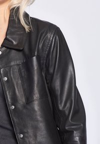 Zwarte leren jas met een kraag, drukknoopsluiting en een borstzak; heeft een gladde textuur en een getailleerde pasvorm.