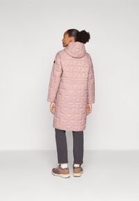 Manteau long matelassé rose clair avec capuche, arborant un motif en diamant, associé à un pantalon gris et des chaussures violettes.