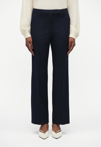 Pantalons large marine fabriqués en tissu lisse, avec un design ajusté comprenant un devant plat et des passants de ceinture. Portés avec des talons blancs.
