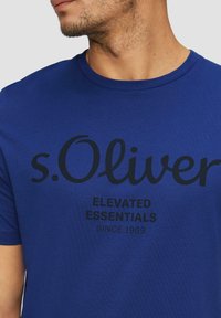Modrá krátkosrstá tričko vyrobené z měkké bavlny, s černým logem "s.Oliver" a textem "ELEVATED ESSENTIALS SINCE 1969" tištěným na přední straně.