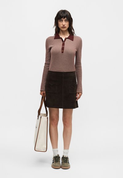 Γυναίκα που φοράει ροζ-μοβ πόλο με μακριά μανίκια και σκοτεινή καφέ φούστα, κρατάει μια λευκή τσάντα tote, με λευκές κάλτσες και καφέ αθλητικά παπούτσια.