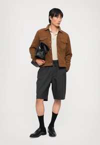 Brązowa zamszowa kurtka, kremowa koszulka polo, szare szorty, czarne mokasyny i skarpetki. Model trzyma czarną kopertówkę. Proste tło.