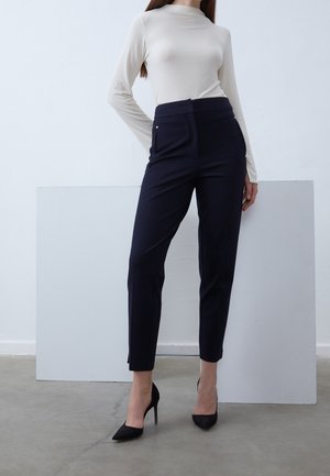 Kobieta ubrana w kremową bluzkę z długim rękawem, granatowe spodnie z wysokim stanem oraz czarne szpilki z ostrym noskiem, stojąca na białym tle.