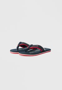 Tumes sinised flip-flops punaste aktsentidega, millel on tekstuuriga jalapadi ja rihmad ning silmapaistev logo taldadel. Vastandlikud valged ja punased äärised.