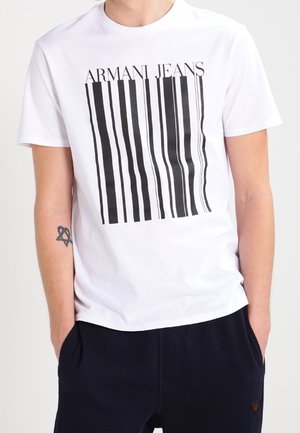 Man in een wit Armani Jeans T-shirt met een zwart verticale streepjescode-design en zwarte broek, handen in de zakken, tatoeage op de linker onderarm.