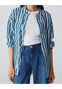 Niebieska i biała koszula w wzór falisty, lekki materiał, zapinana na guziki, z podwiniętymi rękawami; noszona na białej bluzce.