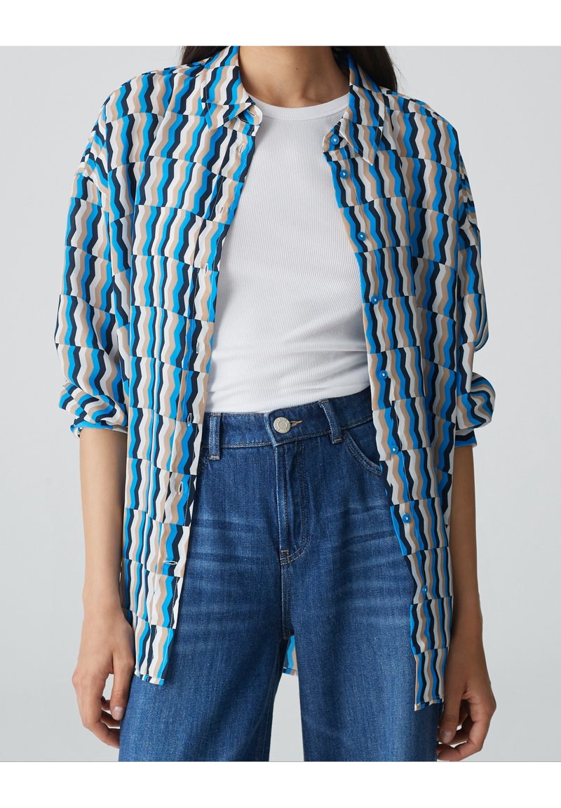 Niebieska i biała koszula w wzór falisty, lekki materiał, zapinana na guziki, z podwiniętymi rękawami; noszona na białej bluzce.