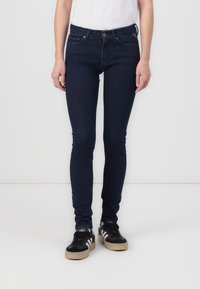 Jeans de mezclilla ajustados, de color azul oscuro, con una textura suave, diseño de cinco bolsillos y trabillas para el cinturón, combinados con zapatillas negras.