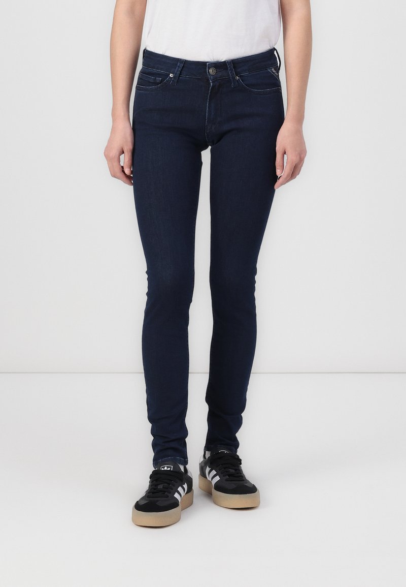 Jeans de mezclilla ajustados, de color azul oscuro, con una textura suave, diseño de cinco bolsillos y trabillas para el cinturón, combinados con zapatillas negras.