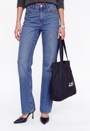 Un mannequin porte un jean bleu droit et des talons noirs, et tient un sac cabas noir JJXX sur un fond de studio blanc.