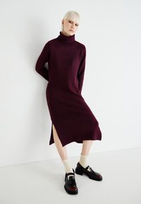 Burgundy knit dress with turtleneck, featuring side slits and a relaxed fit. Paired with black shoes and cream ribbed socks.