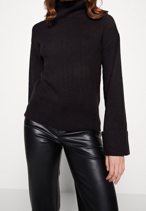 Vrouw die een zwarte coltrui met brede manchetten en een zwarte leren broek draagt, staand tegen een witte achtergrond.