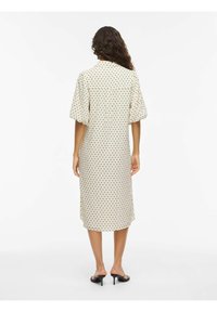 Femme aux longs cheveux bouclés portant une robe beige mi-longue à pois noirs et des sandales à talons noires, debout de dos.