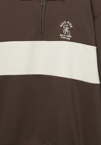 Sweatshirt à blocs de couleurs marron et blanc avec demi-fermeture éclair, présentant un logo de cycliste et le texte "RACE RIDE WILD CARD MARYLAND" sur la poitrine.