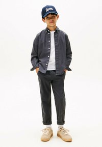 Camisa gris de botones sobre una camiseta blanca, combinada con pantalones grises y zapatillas beige. Gorra azul marino con el logo "Tommy", de pie en un fondo neutro.