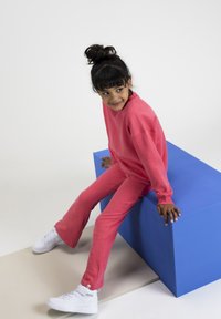 Roze sweatshirt en bijpassende broek gemaakt van zachte stof. Model zit op een blauwe geometrische blok, gekleed in witte sneakers. Glad textuur is zichtbaar.
