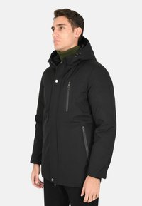Giacca nera con cappuccio, dotata di tasche con zip, con un esterno liscio e un interno caldo e isolato. Presenta una chiusura a scatto e un maglione verde sotto.