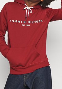 Persona con una sudadera roja de Tommy Hilfiger con cordones blancos y logotipo, combinada con jeans de color azul oscuro, contra un fondo simple.