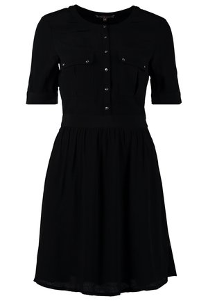 Femme debout, jambes croisées, portant une robe noire mi-longue à boutons et des talons noirs à bride découpée sur fond gris.