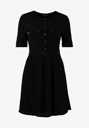Woman standing with crossed legs wearing a black knee-length dress with buttons and black cut-out ankle strap heels on a gray background.