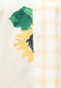 Κίτρινο ηλιοτρόπιο με πράσινα φύλλα τυπωμένο σε κρεμ ύφασμα, ραμμένο δίπλα σε απαλό κίτρινο καρό ύφασμα με αχνό κείμενο "Moschino Jeans".