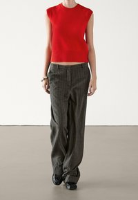 Pull tricoté rouge court avec ourlet côtelé, associé à un pantalon large gris à fines rayures. Le modèle porte des chaussures noires. Design simple et contemporain.