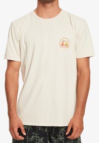 Muž v krémovém tričku Quiksilver s malým kulatým logem zobrazujícím vlnu a horu na levé části hrudi.