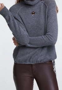 Grijze coltrui met geribbelde manchetten, voorzien van een sequinsterren detail op de borst. Gecombineerd met glanzende bruine leren broeken.